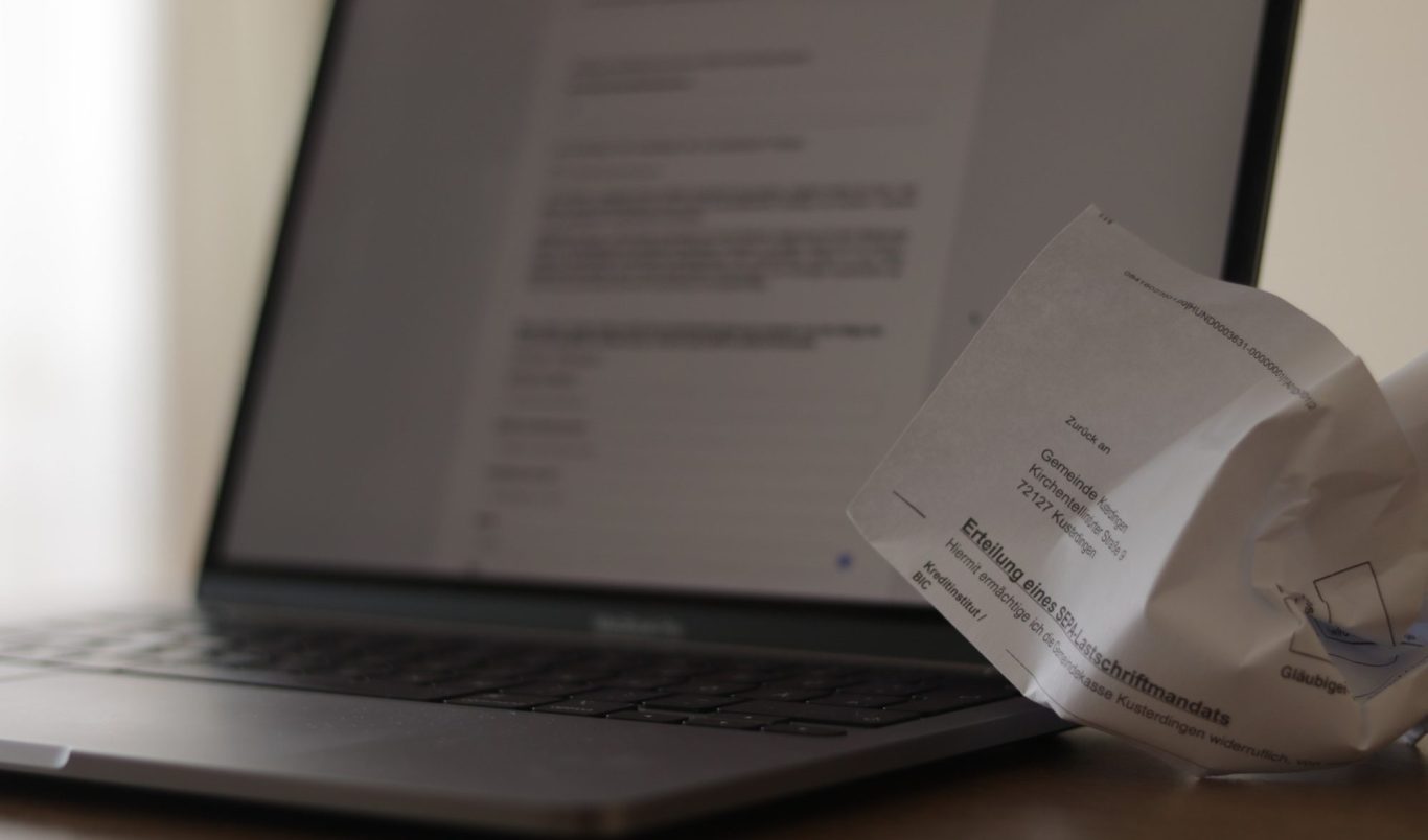 Ein Laptop zeigt eine moderne Benutzeroberfläche für digitale Verwaltungsdienstleistungen. Daneben ein Papierformular der Gemeinde.